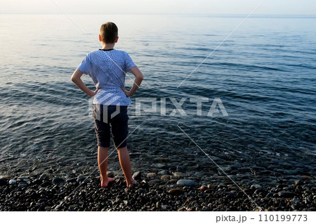 Teen boy standing in sea on pebble beach looking at transparent clear water. 110199773