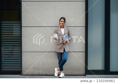 Woman with cocktail and notebook standing near modern building. Woman with cocktail and notebook standing near modern building. 110200861