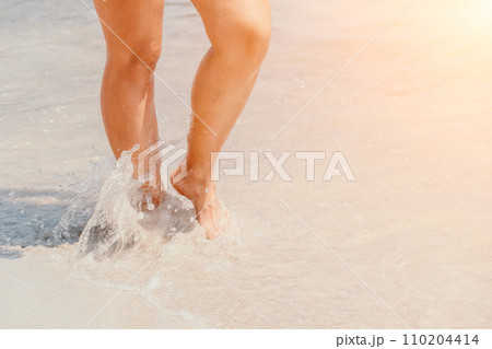 Sea beach travel - woman walking on sand beach leaving footprints in the white sand. Female legs walking along the seaside barefoot, close-up of the tanned legs of a girl coming out of the water. Sea beach travel - woman walking on sand beach leaving footprints in the white sand. Female legs walking along the seaside barefoot, close-up of the tanned legs of a girl coming out of the water. 110204414