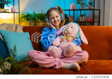 Happy child girl in wireless headphones relaxing sitting on sofa at home listening favorite music 110205836