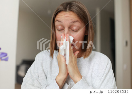 Seasonal allergy. Close up face of allergic young woman uses paper tissues from runny nose, sneezes from flowers pollen at home. Girl with close eyes with flu, itching or cough, allergy rhinitis Seasonal allergy. Close up face of allergic young woman uses paper tissues from runny nose, sneezes from flowers pollen at home. Girl with close eyes with flu, itching or cough, allergy rhinitis 110208533