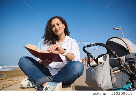 Portrait of authentic mother reading book outdoor, spending nice time on the beach with her baby sleeping in stroller 110209819