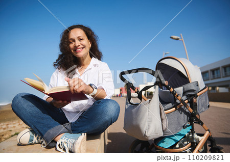 Pretty woman reading book, sitting on a parapet outdoor near a baby stroller, enjoying happy day of her maternity leave Pretty woman reading book, sitting on a parapet outdoor near a baby stroller, enjoying happy day of her maternity leave 110209821