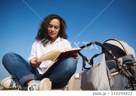 Relaxed young woman reading a book, sitting next to her baby sleeping in the baby pram outdoor 110209822