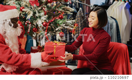 Customer in fashion store talking with actor dressed as Santa Claus, sitting next to Christmas tree during festive promotional event. Client receiving present from worker during holiday season Customer in fashion store talking with actor dressed as Santa Claus, sitting next to Christmas tree during festive promotional event. Client receiving present from worker during holiday season 110210697