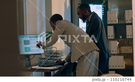 African american businessmen trying to troubleshoot reason for company revenue down from last year, checking numbers in office. Employees looking over financial charts during nightshift 110210744