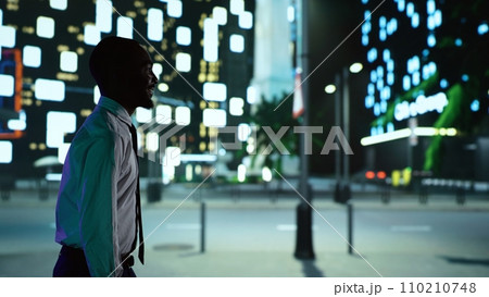 African american person on night walk downtown under streetlights, admiring modern office buildings from illuminated sidewalk. Confident man enjoying nighttime promenade around town. 110210748