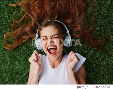 Happy teenage girl listening to music with headphones lying on the green lawn grass in the park, summer vacation 110212218