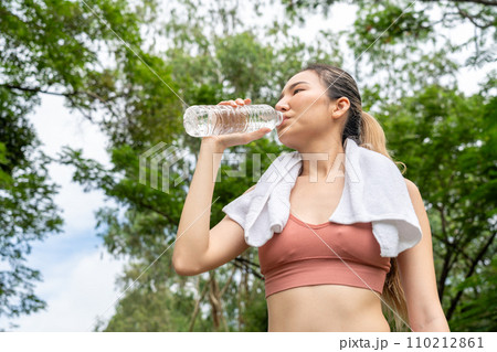 Young Asian woman in fitness clothes stops to drink water from her plastic water bottle during her morning exercise run at  running track of a local park 110212861