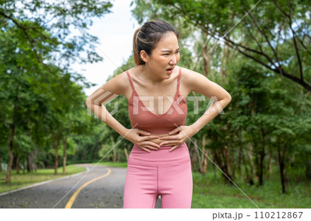 Young atractive Asian woman in fitness clothes with stomach pain putting her hands over her stomach while standing at running track of a local park 110212867