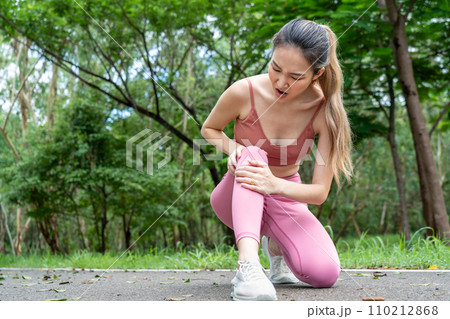 Young atractive Asian woman in fitness clothes with knee pain putting her hands over her troubled right knee while sitting down at running track of a local park 110212868