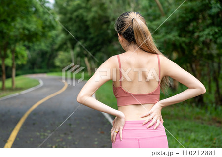 Back view of a long hair woman with back pain putting her hands over her trouble lower back while standing at running track of a local park Back view of a long hair woman with back pain putting her hands over her trouble lower back while standing at running track of a local park 110212881