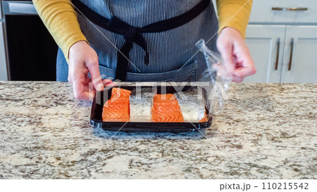Woman is opening a store package containing raw salmon while preparing to bake teriyaki salmon in a modern white kitchen. Woman is opening a store package containing raw salmon while preparing to bake teriyaki salmon in a modern white kitchen. 110215542