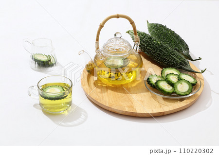 On the wooden tray there is a transparent teapot, slices of bitter melon and fresh bitter melon fruit, next to a cup of tea and a glass vase. Minimalist white background for advertising. 110220302