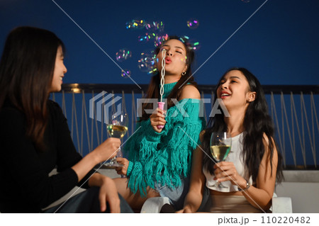 Group of young people having fun at outdoor rooftop party with drinks and blowing soap bubbles. 110220482
