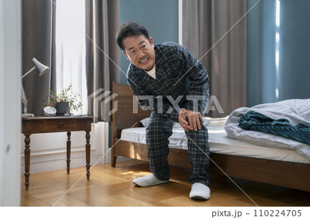 ベッドのそばで膝に手をあてるパジャマ姿の50代半ばの男性 ベッドのそばで膝に手をあてるパジャマ姿の50代半ばの男性 110224705