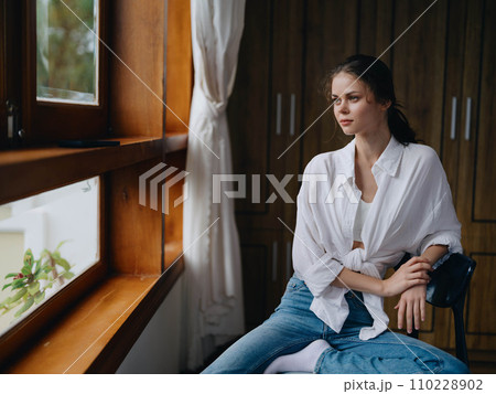 Woman sitting at home by a wooden window with a smile on a chair in homemade comfortable clothes and looking at the landscape, spring mood, women's day, rest on the weekend. 110228902