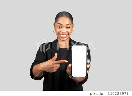 Smiling young african american woman in formal suit point finger at smartphone with empty screen 110228971
