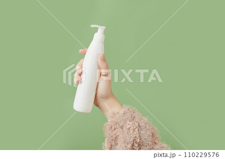 A womans hand in a sweater holds a bottle of cream on a green background. Self care beauty treatment A womans hand in a sweater holds a bottle of cream on a green background. Self care beauty treatment 110229576