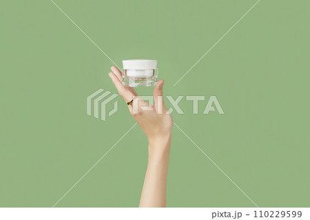 A womans hand holding a white cosmetic jar on a green background A womans hand holding a white cosmetic jar on a green background 110229599