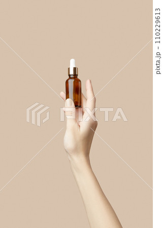 A womans hand holding a brown bottle of serum on a beige background. Body care A womans hand holding a brown bottle of serum on a beige background. Body care 110229613