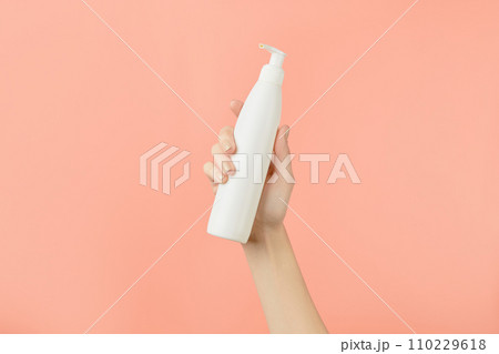 A womans hand holding a white cosmetic bottle on a peach background. Beauty treatment concept A womans hand holding a white cosmetic bottle on a peach background. Beauty treatment concept 110229618
