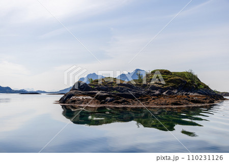 Tranquil islet with moss-covered outcrops mirrored in calm waters under a hazy Lofoten sky Tranquil islet with moss-covered outcrops mirrored in calm waters under a hazy Lofoten sky 110231126