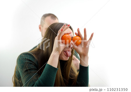 young girl put Tangerines to eyes and pulled out tongue stuck out and she twists and makes faces in the background a man on a white background a green sweater long hair joke fooling around grimacing young girl put Tangerines to eyes and pulled out tongue stuck out and she twists and makes faces in the background a man on a white background a green sweater long hair joke fooling around grimacing 110231350