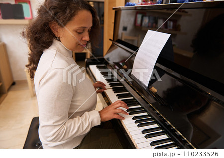 Beautiful multi ethnic young woman, pianist practicing music lesson at home, sitting alone at piano and playing melody Beautiful multi ethnic young woman, pianist practicing music lesson at home, sitting alone at piano and playing melody 110233526