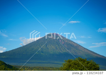 Panorama of Mount Agung and rice fields on the island of Bali. View of the mountain against a background of palm trees and a cornfield. Panorama of Agung volcano covered with clouds on a sunny day. 110234107