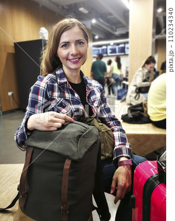 Young woman in shirt waiting for her transfer, with her backpack and luggage near her, looking into camera, smiling, blurred station in background Young woman in shirt waiting for her transfer, with her backpack and luggage near her, looking into camera, smiling, blurred station in background 110234340