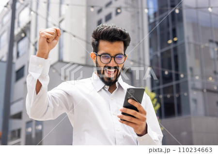 Young successful hispanic man in casual clothes outside office building received online win notification, man celebrating victory triumph, using app on phone, good news in businessman 110234663