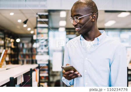 Black man walking along bookcases with smartphone and smiling Black man walking along bookcases with smartphone and smiling 110234776