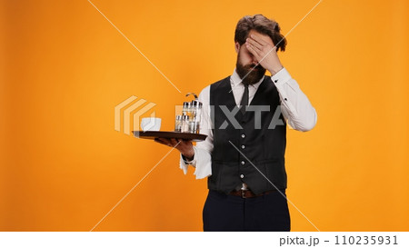 Unhappy butler deals with painful migraine, rubbing temples and feeling overworked at catering job on camera. Young worker in pain with headache suffering from burnout and stress, yellow studio. 110235931