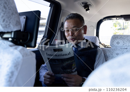 タクシーの後部座席で英字新聞を読む中高年ビジネスマン 110236204