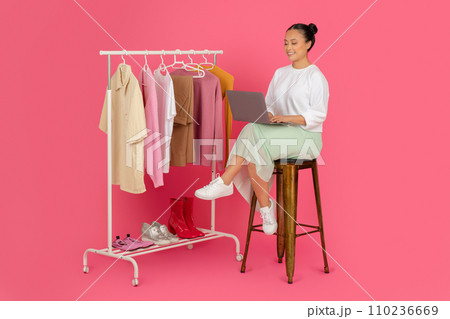 Beautiful asian woman sitting with laptop near clothing rack, managing online boutique Beautiful asian woman sitting with laptop near clothing rack, managing online boutique 110236669