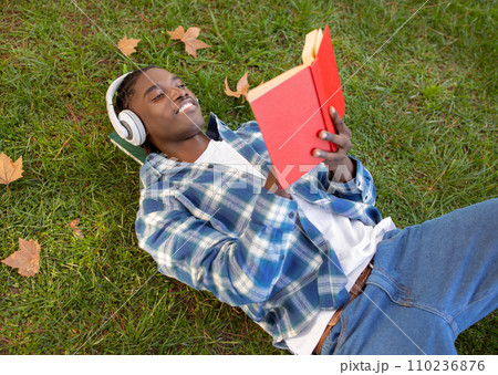 Relaxed black young man enjoying book with headphones lying outside 110236876