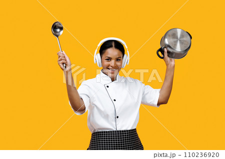 Black female chef enjoying music in headphones while holding ladle and pot 110236920
