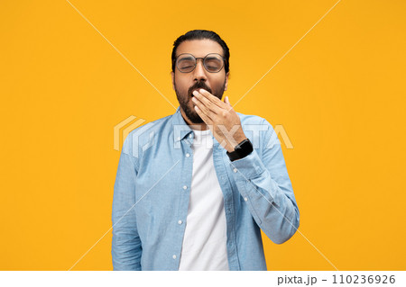 Sleepy or bored man in a light blue shirt, covering his mouth while yawning 110236926