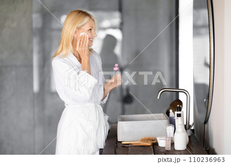 Smiling mature woman wearing white bathrobe applying refreshing facial toner in bathroom Smiling mature woman wearing white bathrobe applying refreshing facial toner in bathroom 110236963