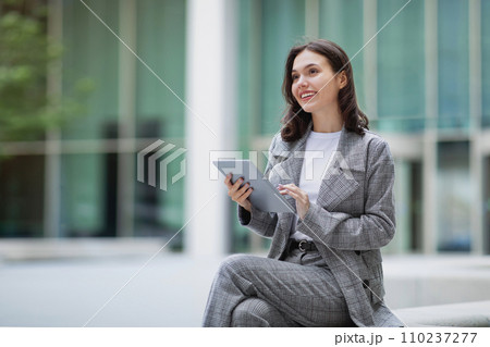 Cheerful young businesswoman working on tablet computer while sitting outdoors 110237277