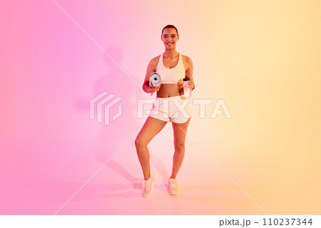 A smiling woman with a shaved head holds a yoga mat and a water bottle, ready for her fitness routine 110237344