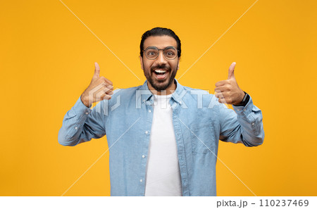 Enthusiastic man in a blue denim shirt giving two thumbs up, wide-eyed and smiling 110237469