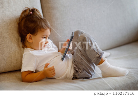 Cute toddler girl using smartphone while sitting on cozy sofa Cute toddler girl using smartphone while sitting on cozy sofa 110237516