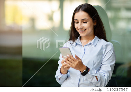 A young businesswoman checks her smartphone with a content smile, wearing a smart watch A young businesswoman checks her smartphone with a content smile, wearing a smart watch 110237547