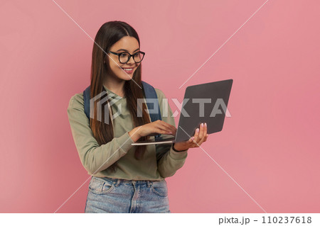 Smiling teenage girl wearing eyeglasses and backpack looking at laptop screen Smiling teenage girl wearing eyeglasses and backpack looking at laptop screen 110237618