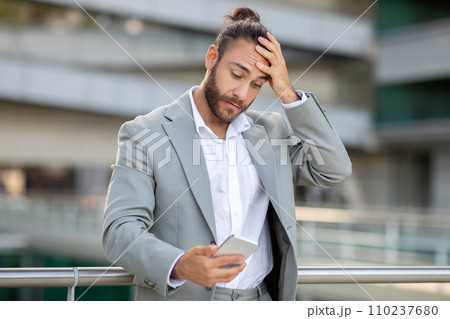 Concerned young businessman reading message on his smartphone while standing outdoors 110237680