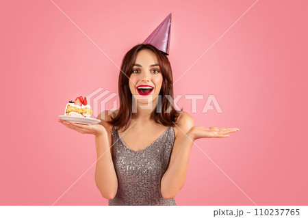 Excited woman in a glittery dress with a party hat presenting a delicious piece of cake 110237765