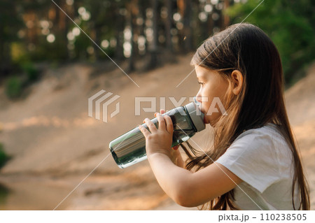 Little girl with dark hair drinking water from bottle sitting outdoor. Active kid resting on sandy beach in nature. Little girl with dark hair drinking water from bottle sitting outdoor. Active kid resting on sandy beach in nature. 110238505