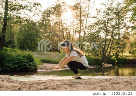 Little athlete doing squats on green mat on sandy beach in front of lake. Focused child working out at sunset outdoor. Little athlete doing squats on green mat on sandy beach in front of lake. Focused child working out at sunset outdoor. 110238522